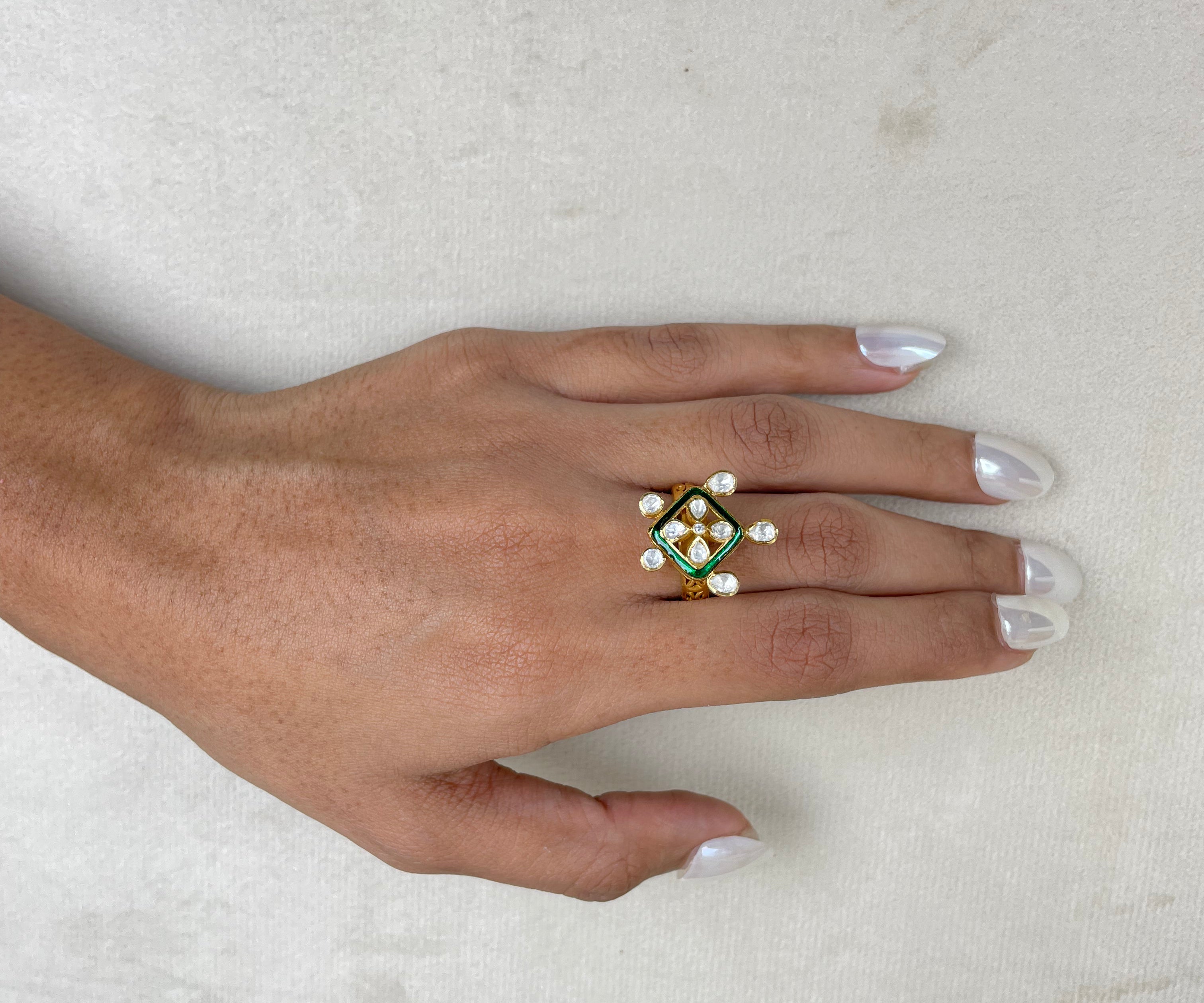 A hand with white manicured nails wearing a gold ring decorated with green enamel, white pearls, and a central clear stone, resting on a textured off-white surface.