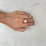 A hand with white manicured nails wearing a gold ring with a large white pearl-like center and red floral accents, resting on a textured light gray surface.