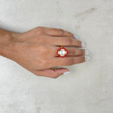 A hand with white manicured nails wearing a gold ring with a large white pearl-like center and red floral accents, resting on a textured light gray surface.