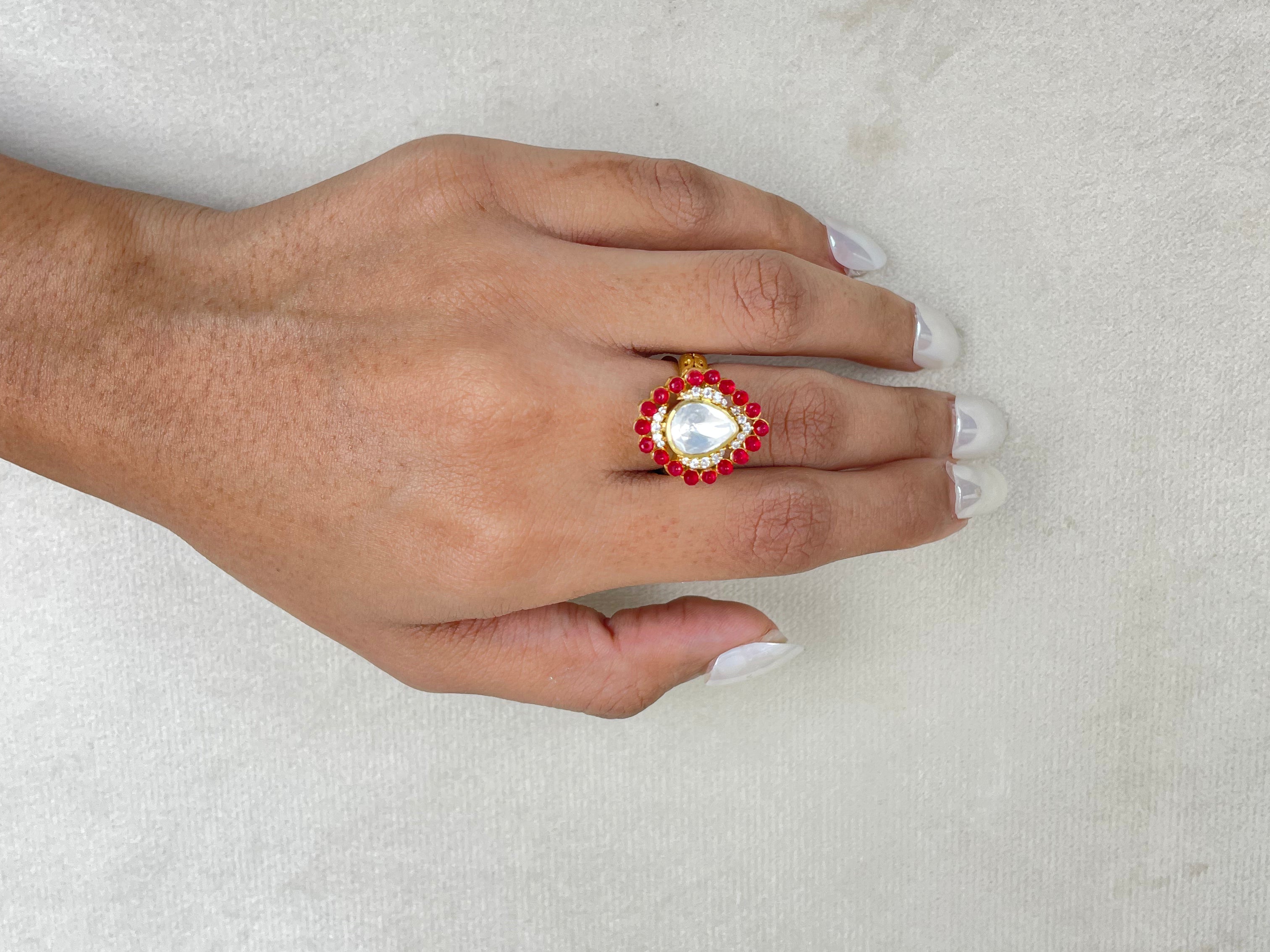 A hand with white manicured nails wearing a gold ring with a large white pearl-like center and red floral accents, resting on a textured light gray surface.