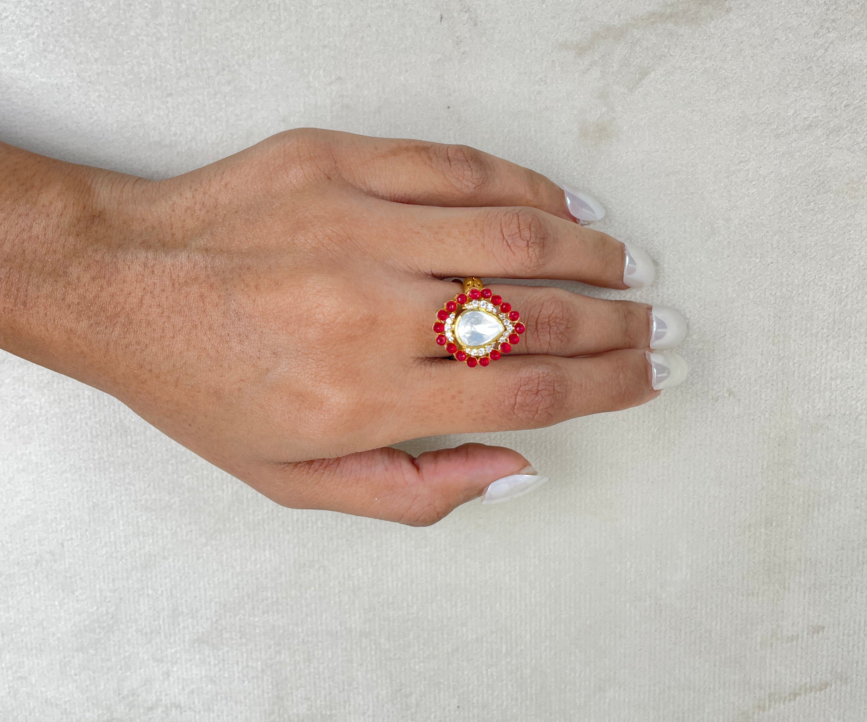 A hand with white manicured nails wearing a gold ring with a large white pearl-like center and red floral accents, resting on a textured light gray surface.