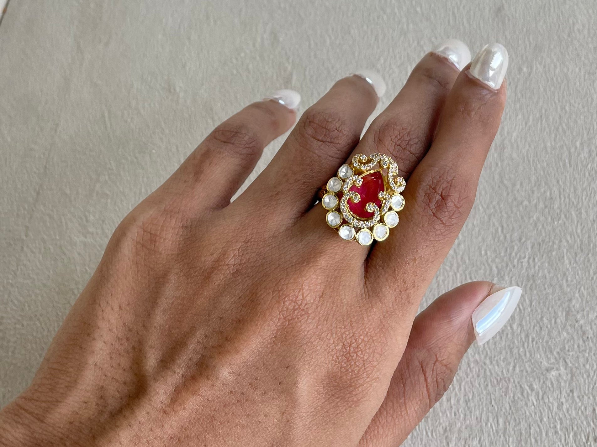 A close-up of a hand wearing a gold ring with a red oval center and a border of small white stones, set against a neutral textured background.