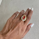 A close-up of a hand wearing a gold ring with a red oval center and a border of small white stones, set against a neutral textured background.