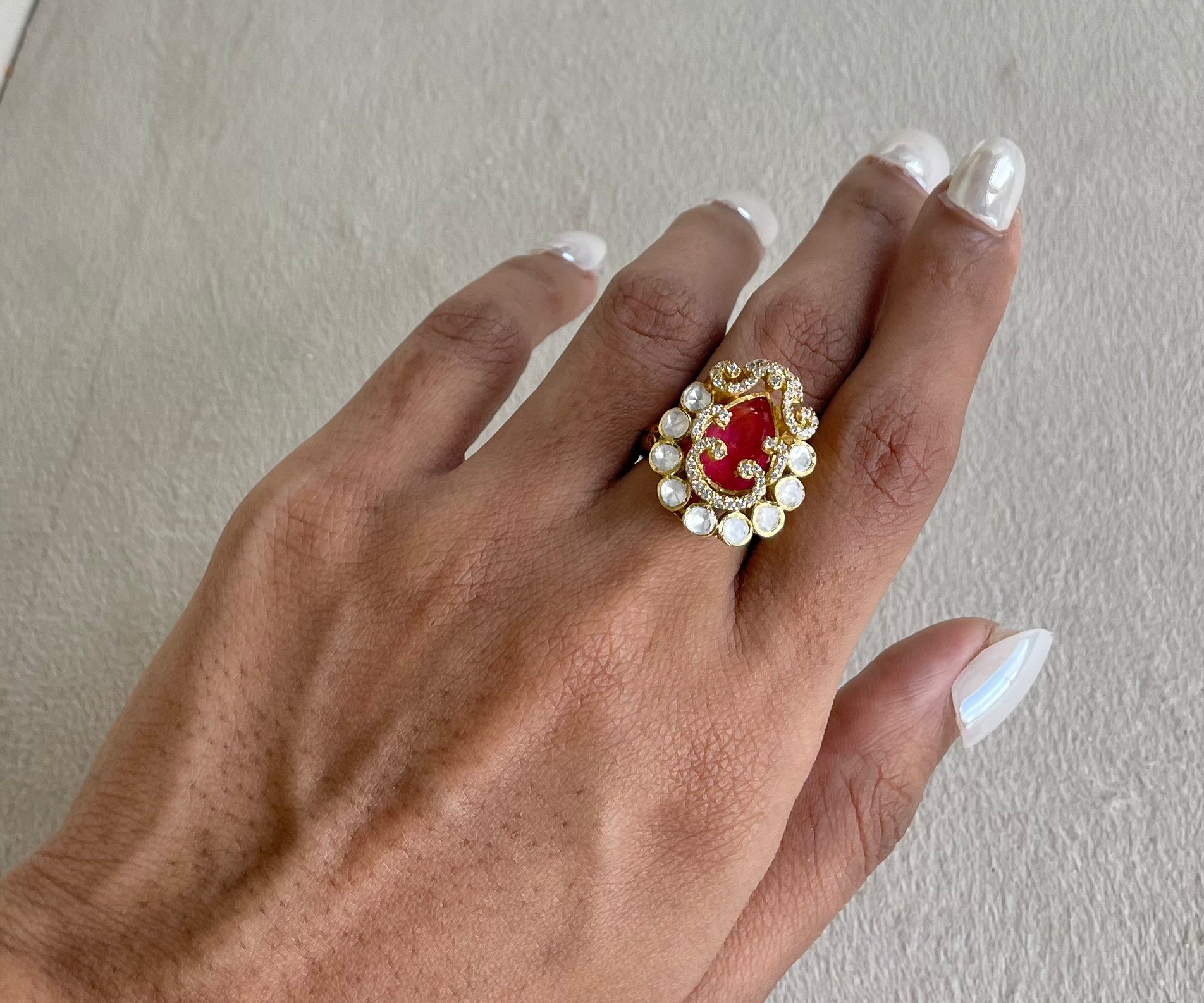 A close-up of a hand wearing a gold ring with a red oval center and a border of small white stones, set against a neutral textured background.