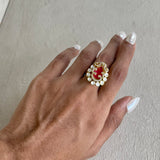A close-up of a hand wearing a gold ring with a red oval center and a border of small white stones, set against a neutral textured background.