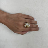 A close-up of a hand with white manicured nails wearing a large gold ring decorated with clear crystals and a central pearl, set against a textured off-white background.