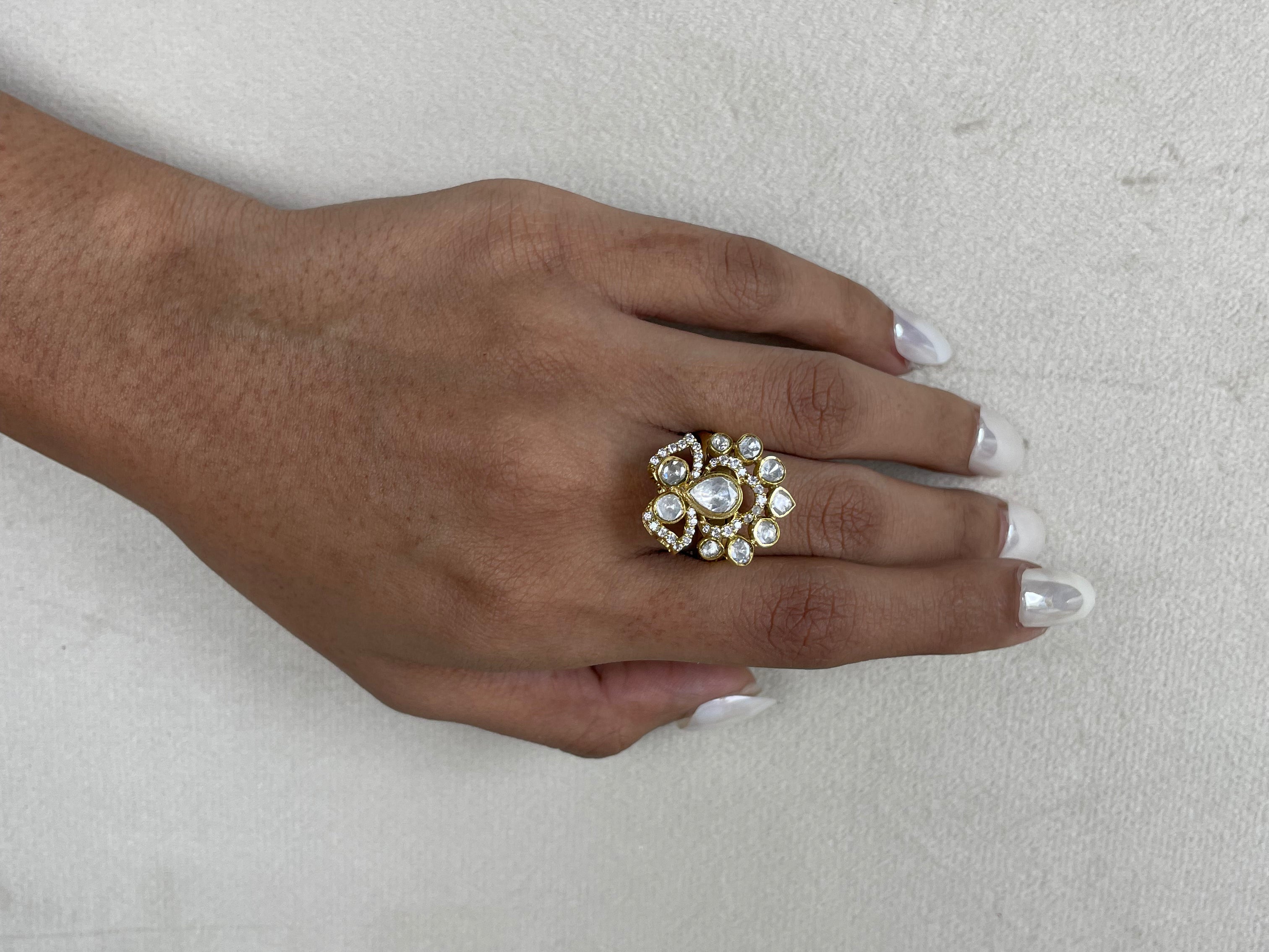 A close-up of a hand with white manicured nails wearing a large gold ring decorated with clear crystals and a central pearl, set against a textured off-white background.