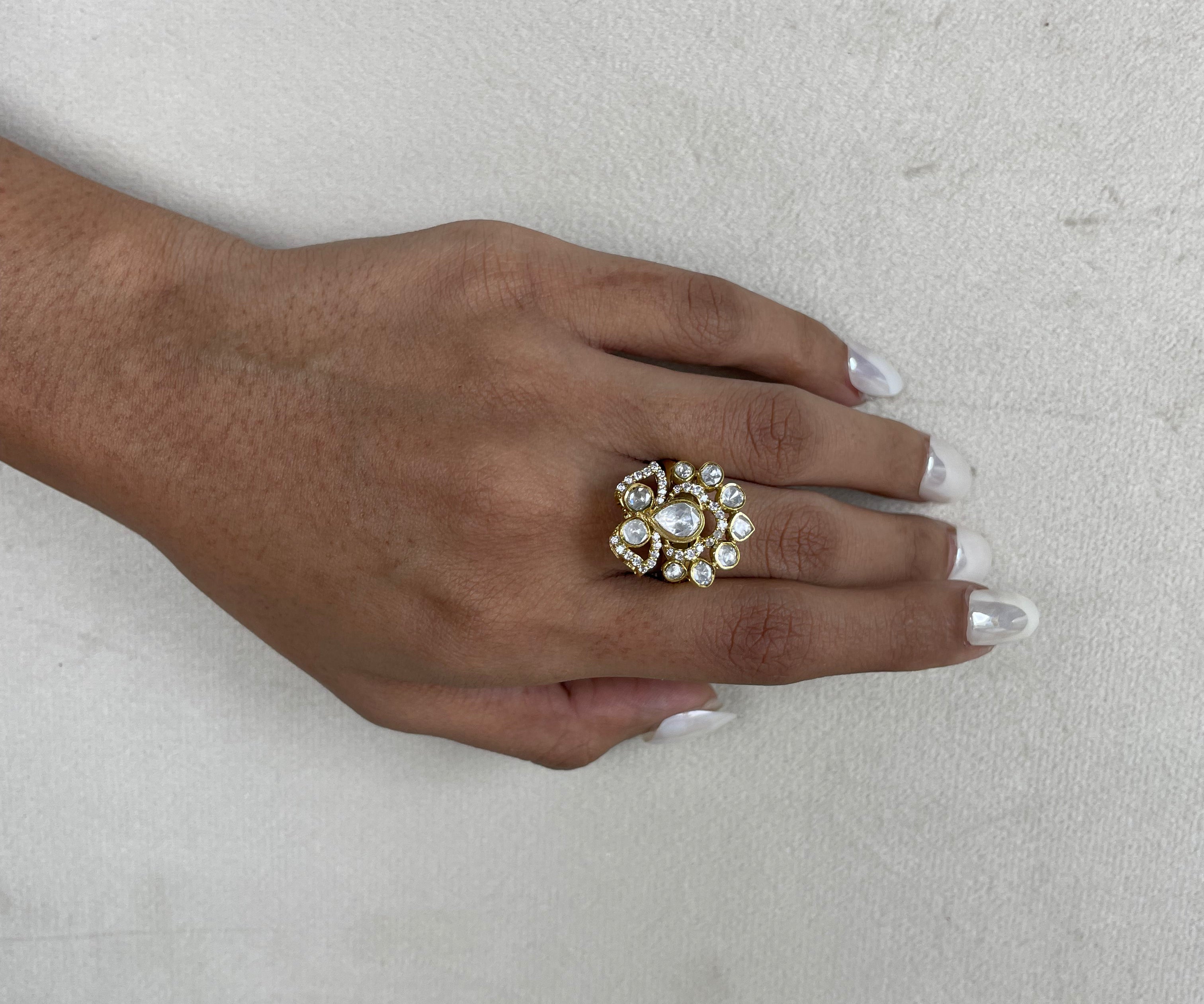 A close-up of a hand with white manicured nails wearing a large gold ring decorated with clear crystals and a central pearl, set against a textured off-white background.