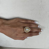 A close-up of a hand with silver manicured nails wearing a gold ring featuring a teardrop-shaped clear gemstone surrounded by a halo of small clear stones.