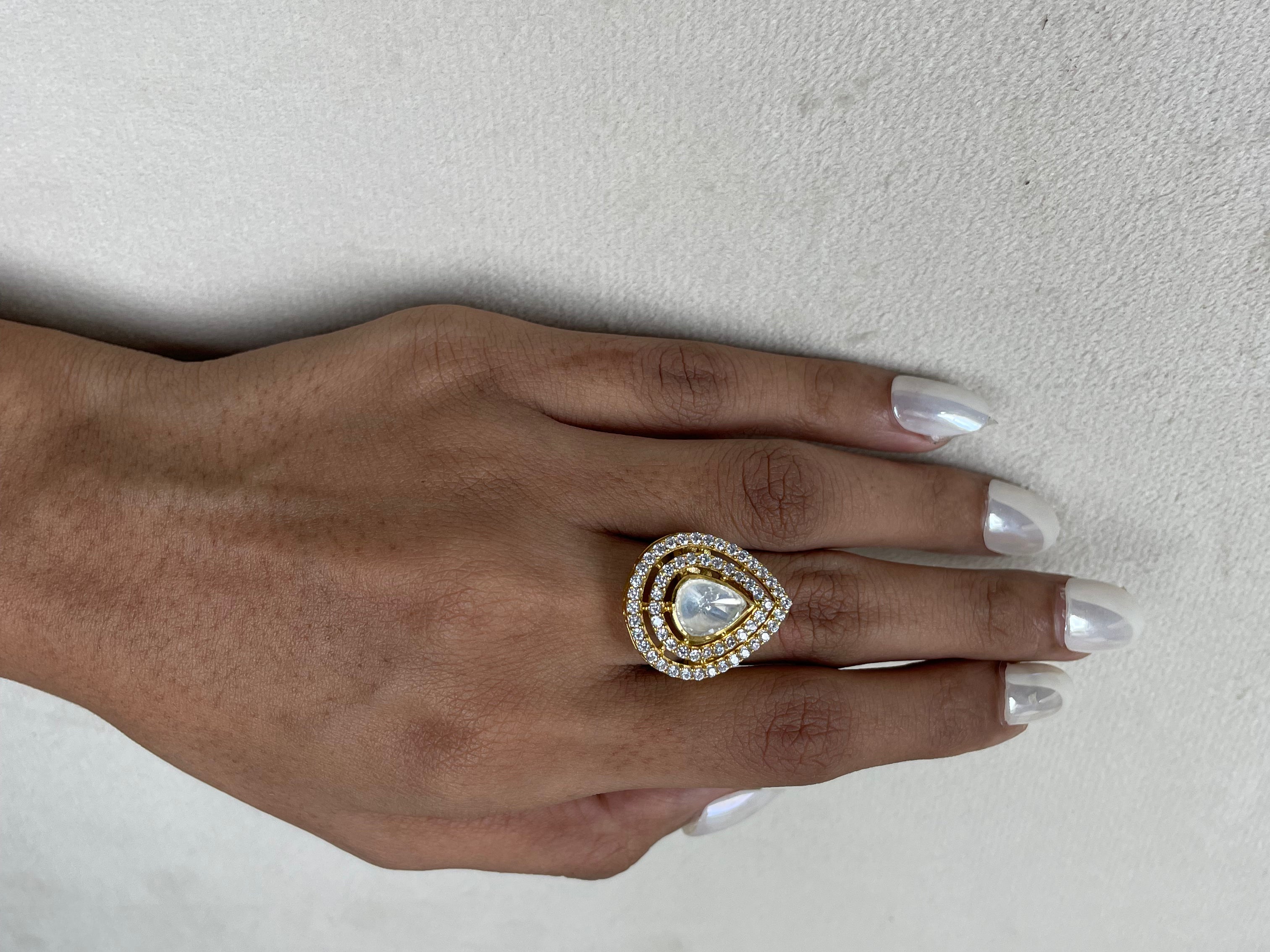 A close-up of a hand with silver manicured nails wearing a gold ring featuring a teardrop-shaped clear gemstone surrounded by a halo of small clear stones.