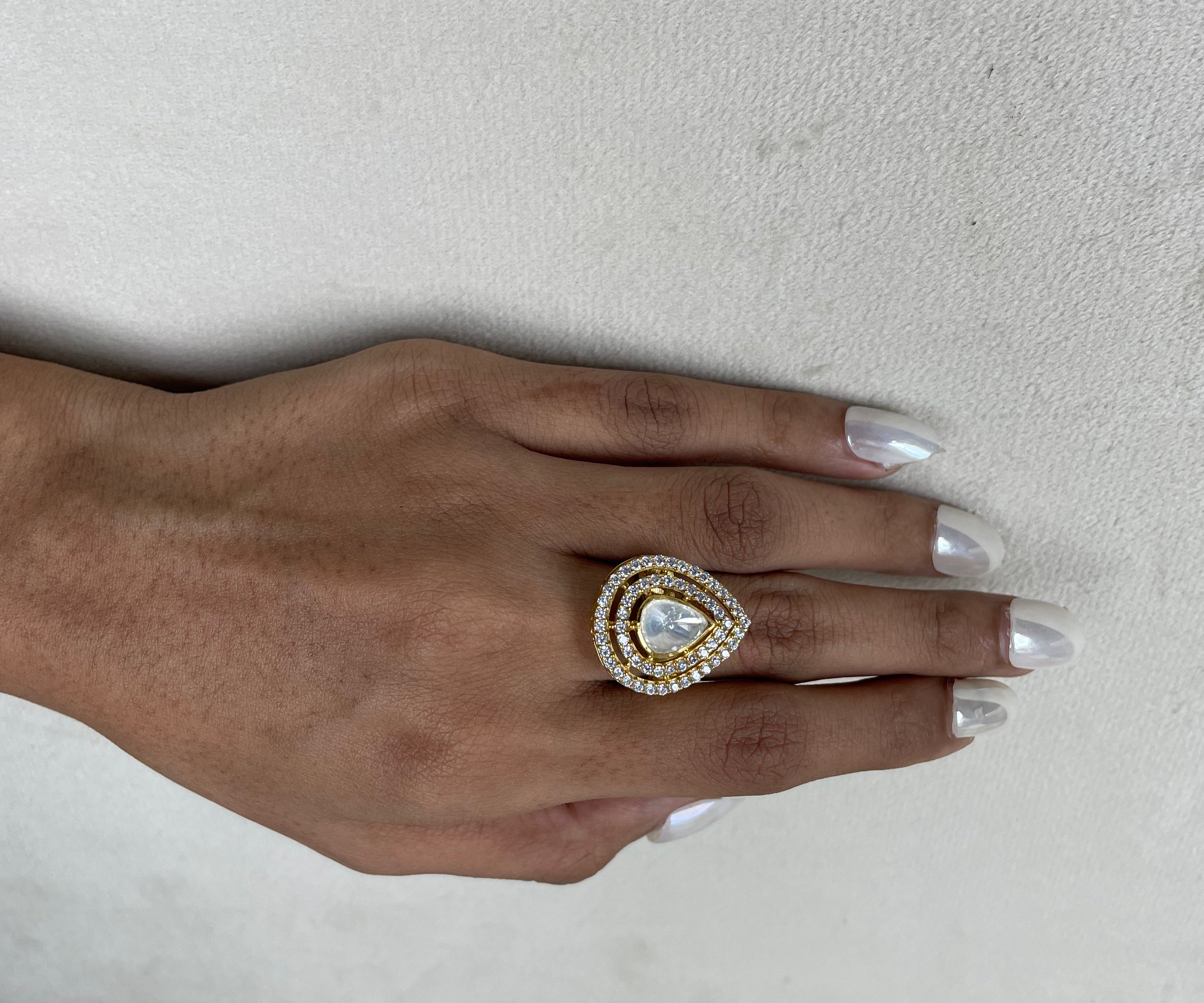 A close-up of a hand with silver manicured nails wearing a gold ring featuring a teardrop-shaped clear gemstone surrounded by a halo of small clear stones.