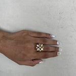 A gold-plated ring with clear stones arranged in a floral or starburst pattern, worn on a person's hand with silver manicured nails, set against a neutral textured light gray background.