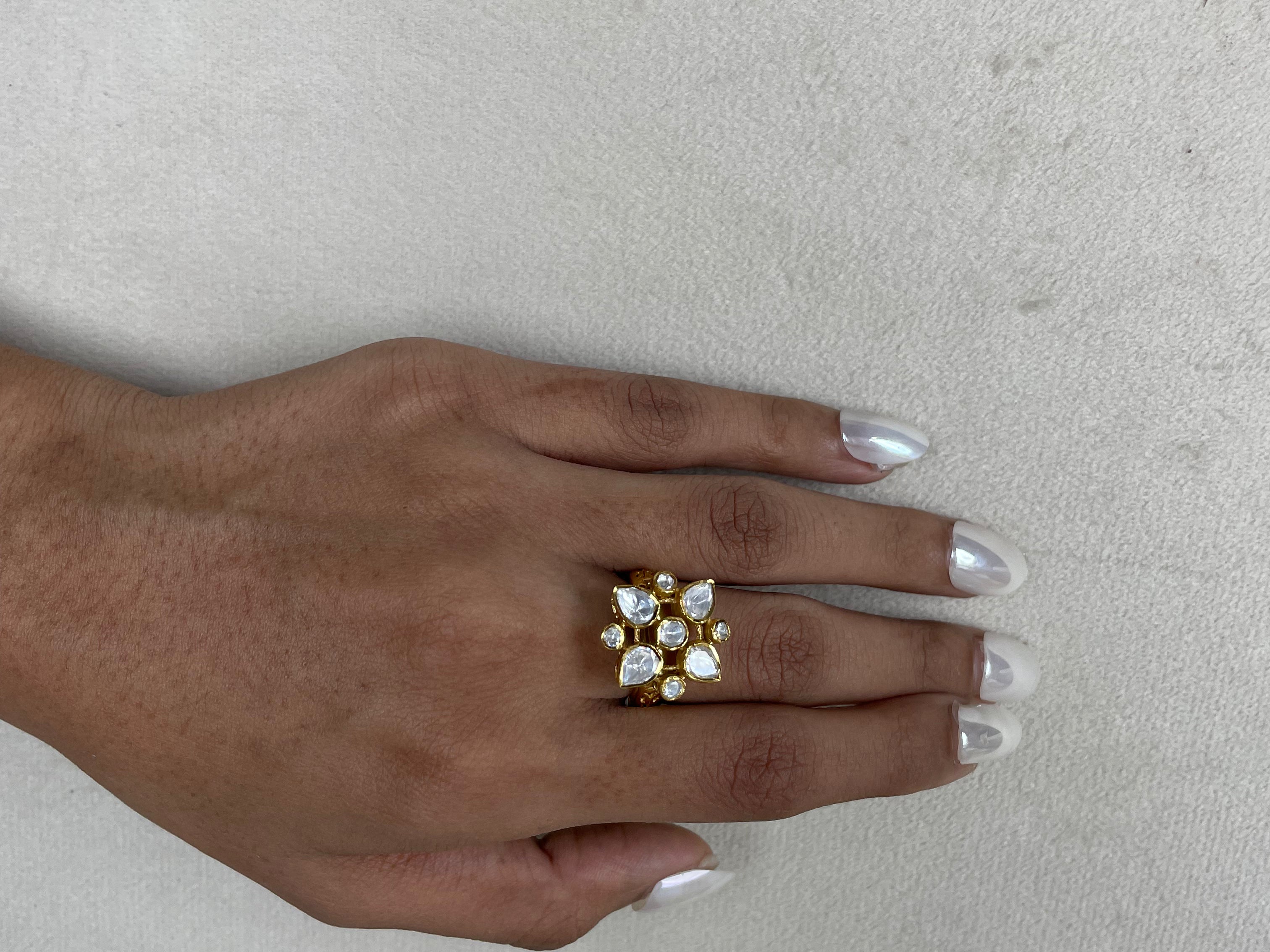 A gold-plated ring with clear stones arranged in a floral or starburst pattern, worn on a person's hand with silver manicured nails, set against a neutral textured light gray background.