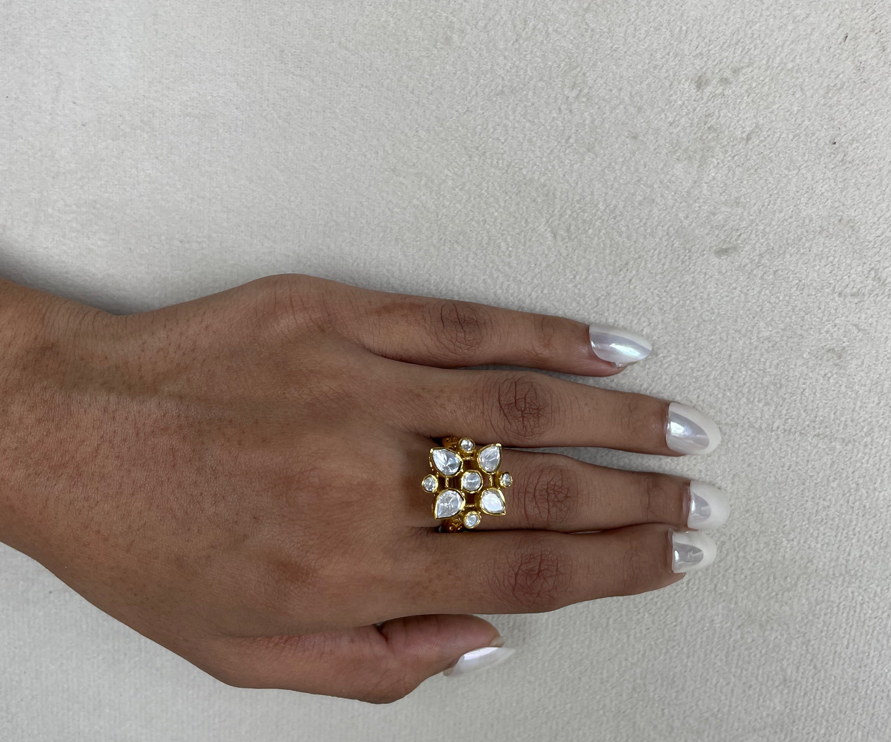 A gold-plated ring with clear stones arranged in a floral or starburst pattern, worn on a person's hand with silver manicured nails, set against a neutral textured light gray background.
