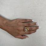 A gold ring with a triangular crystal cluster setting worn on a person's ring finger, shown against a textured light gray background.
