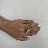 A gold ring with a triangular crystal cluster setting worn on a person's ring finger, shown against a textured light gray background.