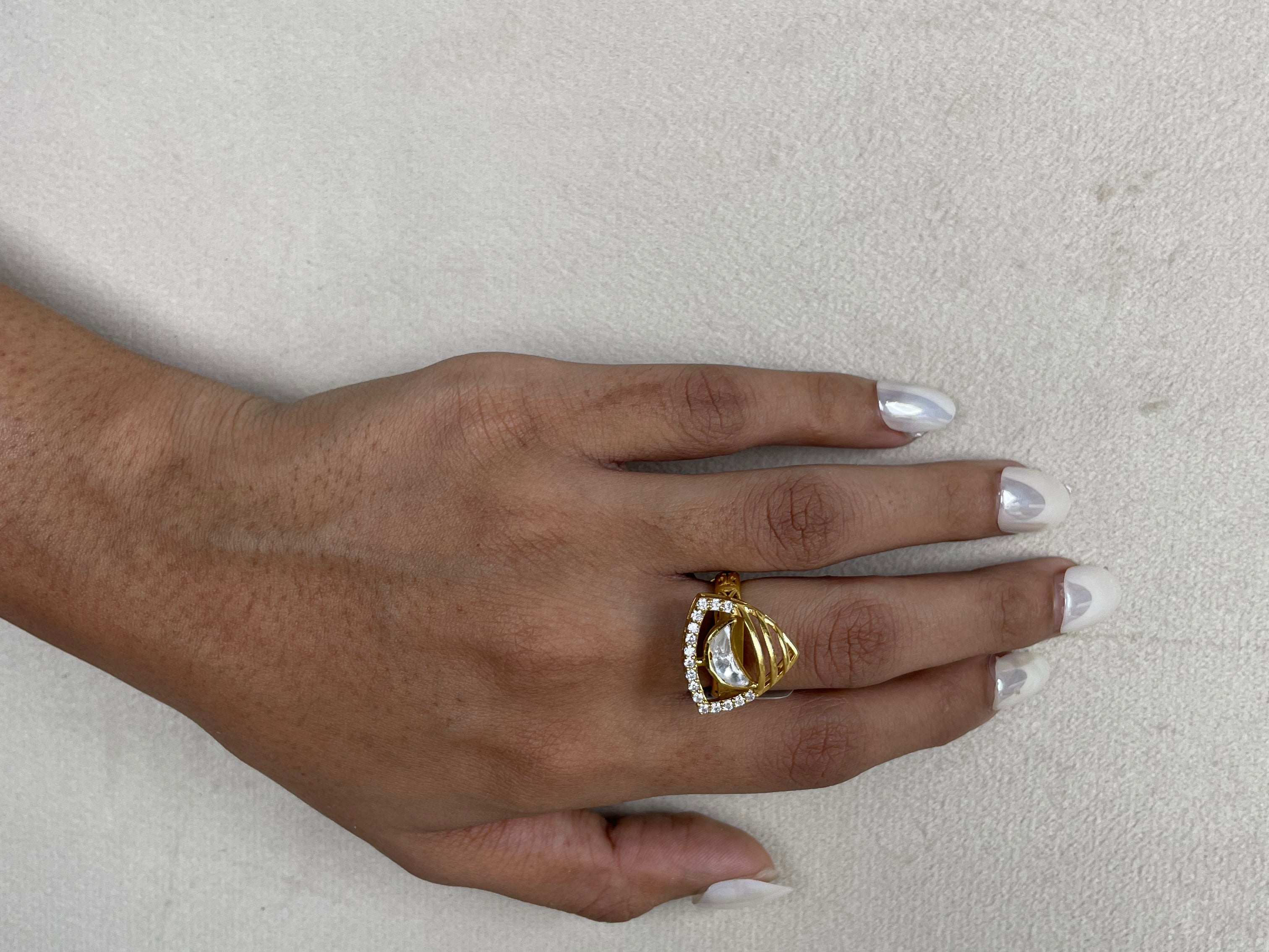 A gold ring with a triangular crystal cluster setting worn on a person's ring finger, shown against a textured light gray background.