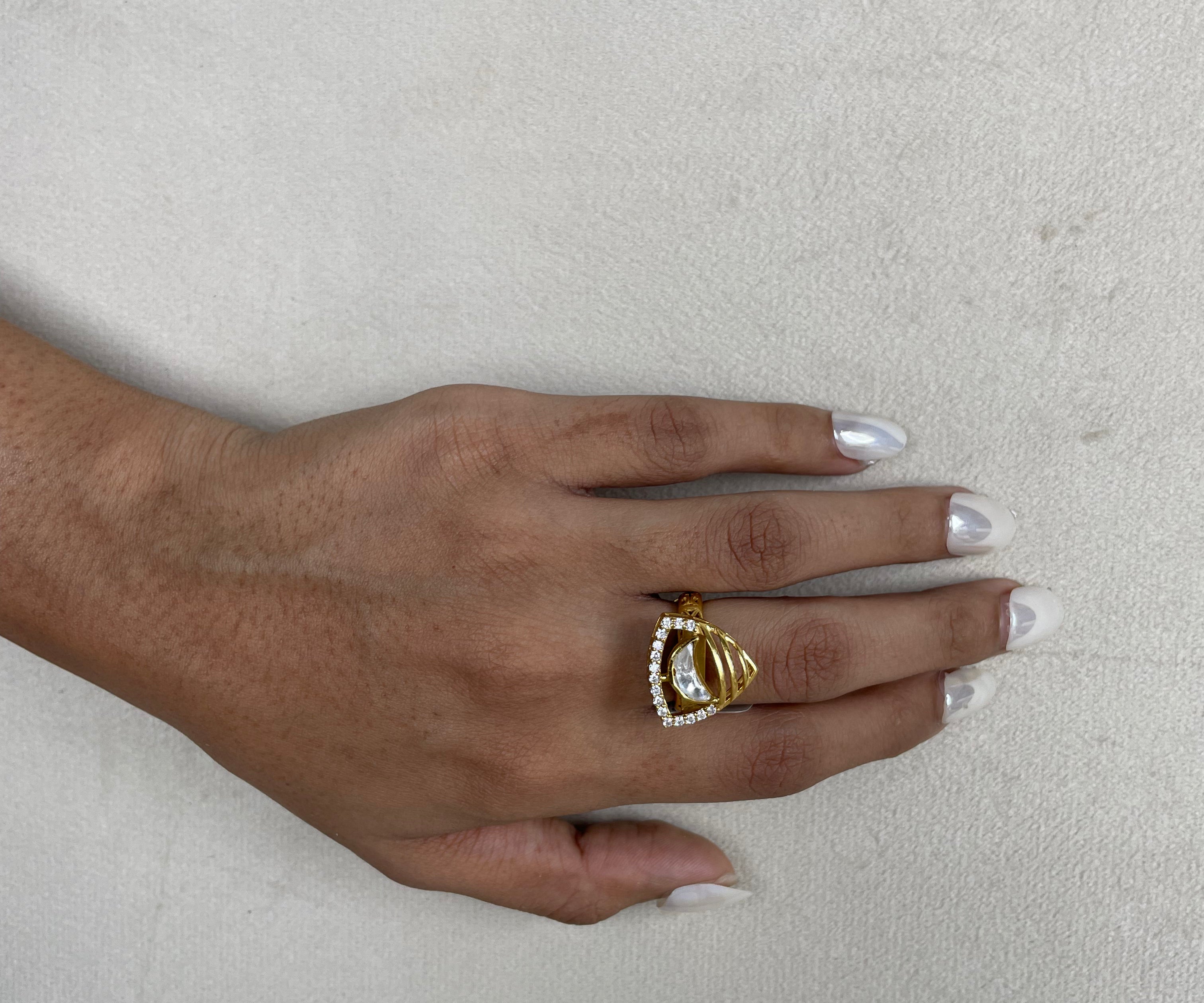 A gold ring with a triangular crystal cluster setting worn on a person's ring finger, shown against a textured light gray background.