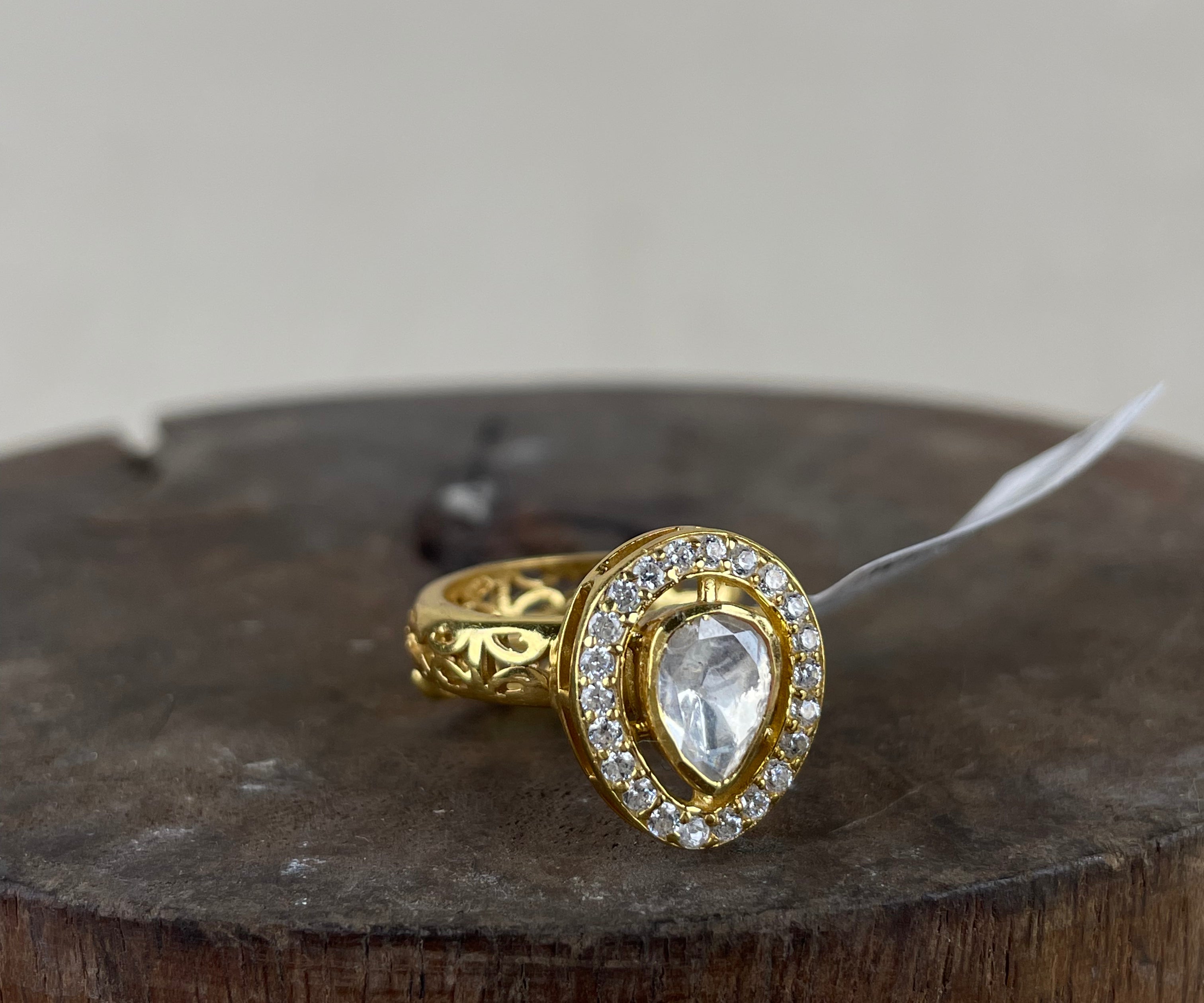A silver gold-plated ring featuring a pear-cut clear gemstone surrounded by a halo of small clear stones, displayed on a rustic wooden block against a neutral beige background.
