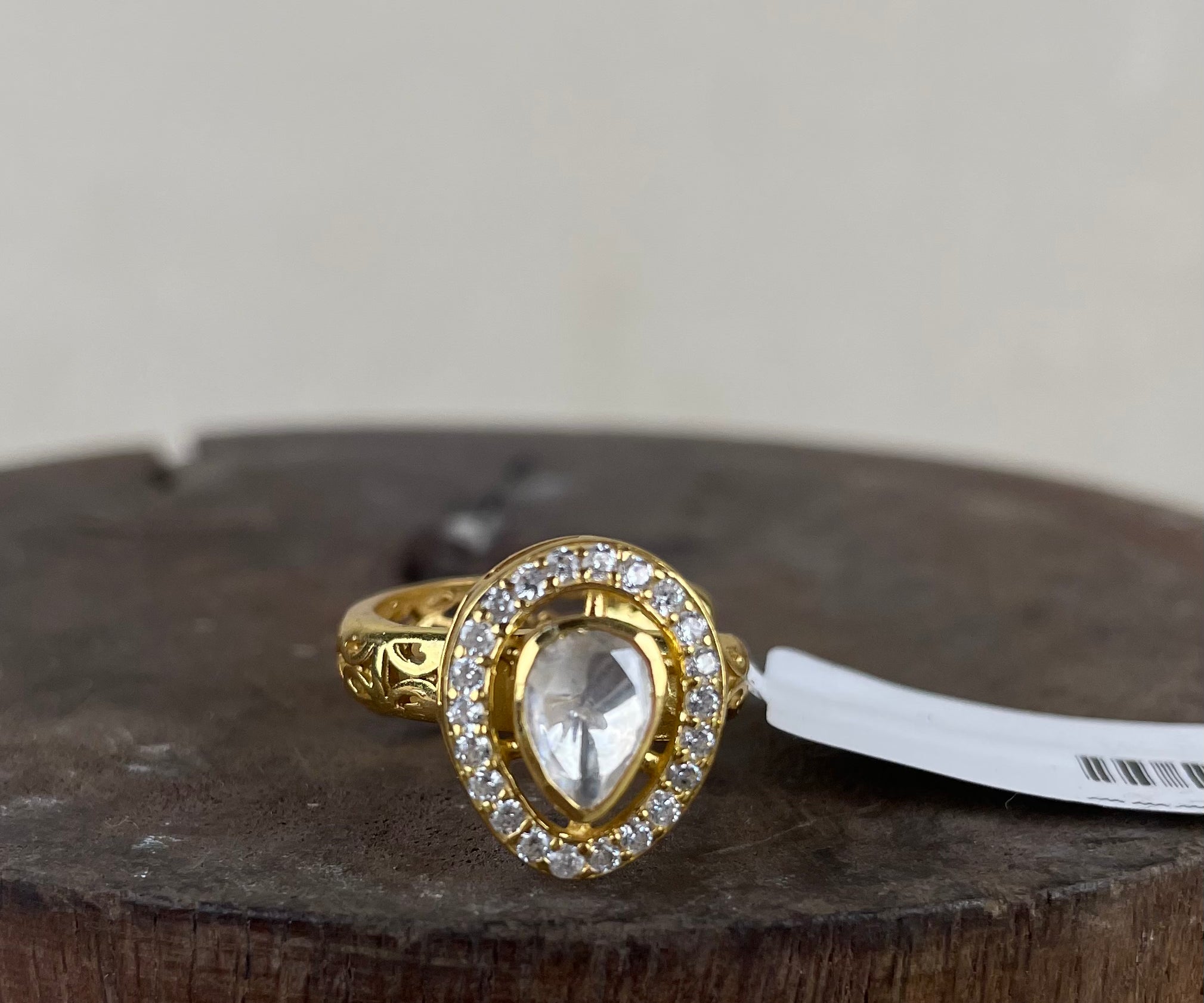 A gold ring featuring a pear-cut clear center stone surrounded by a halo of small clear stones, displayed on a rustic wooden surface with a white tag attached.