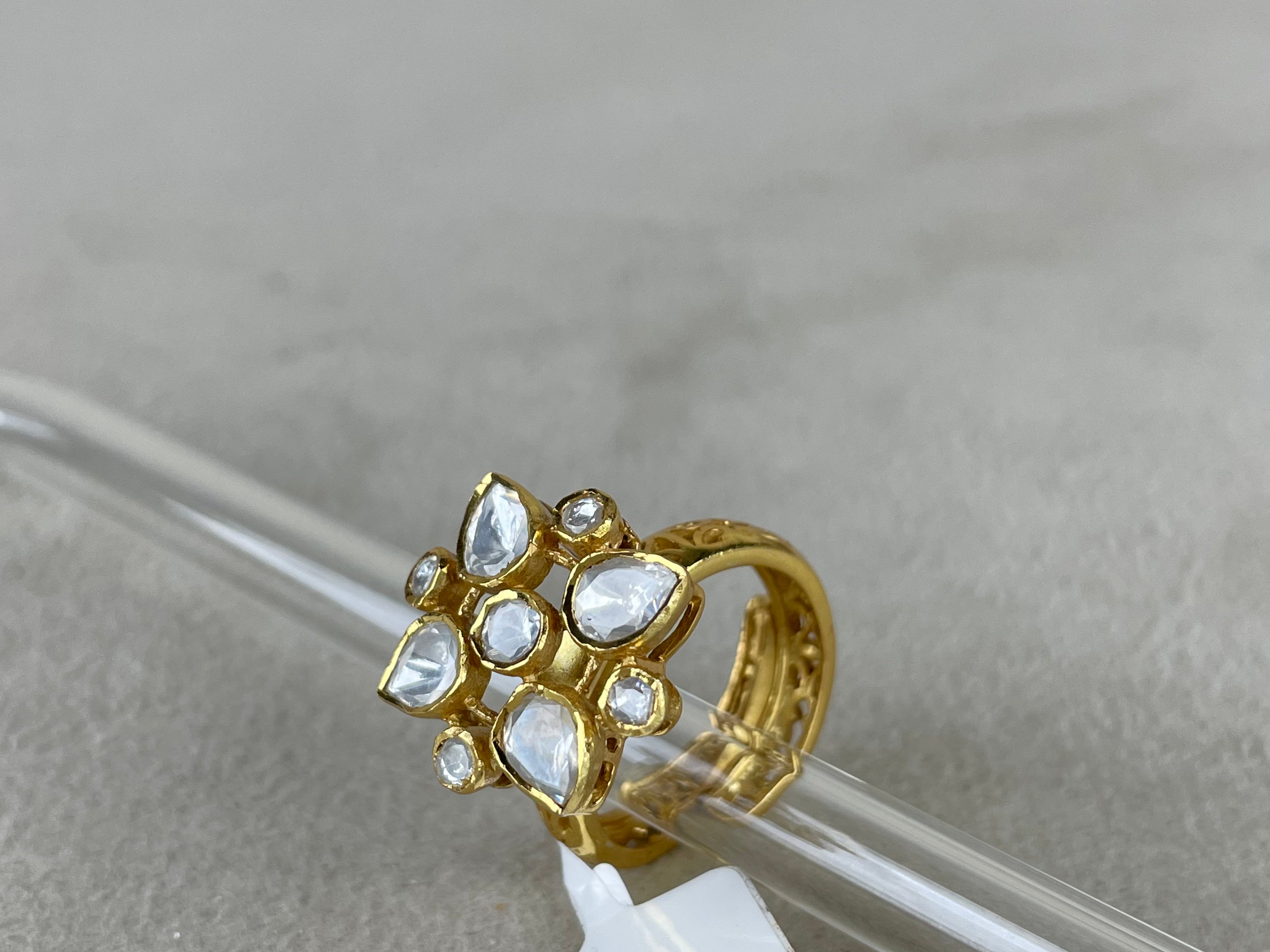 A gold-plated ring with multiple clear teardrop-shaped stones, resting on a clear glass rod with a white product tag attached, set against a neutral gray background.