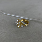A gold-plated ring with a cluster of clear gemstones, resting on a textured gray surface with a clear glass rod in the background.