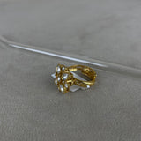 A gold-plated ring with a cluster of clear gemstones, resting on a textured gray surface with a clear glass rod in the background.