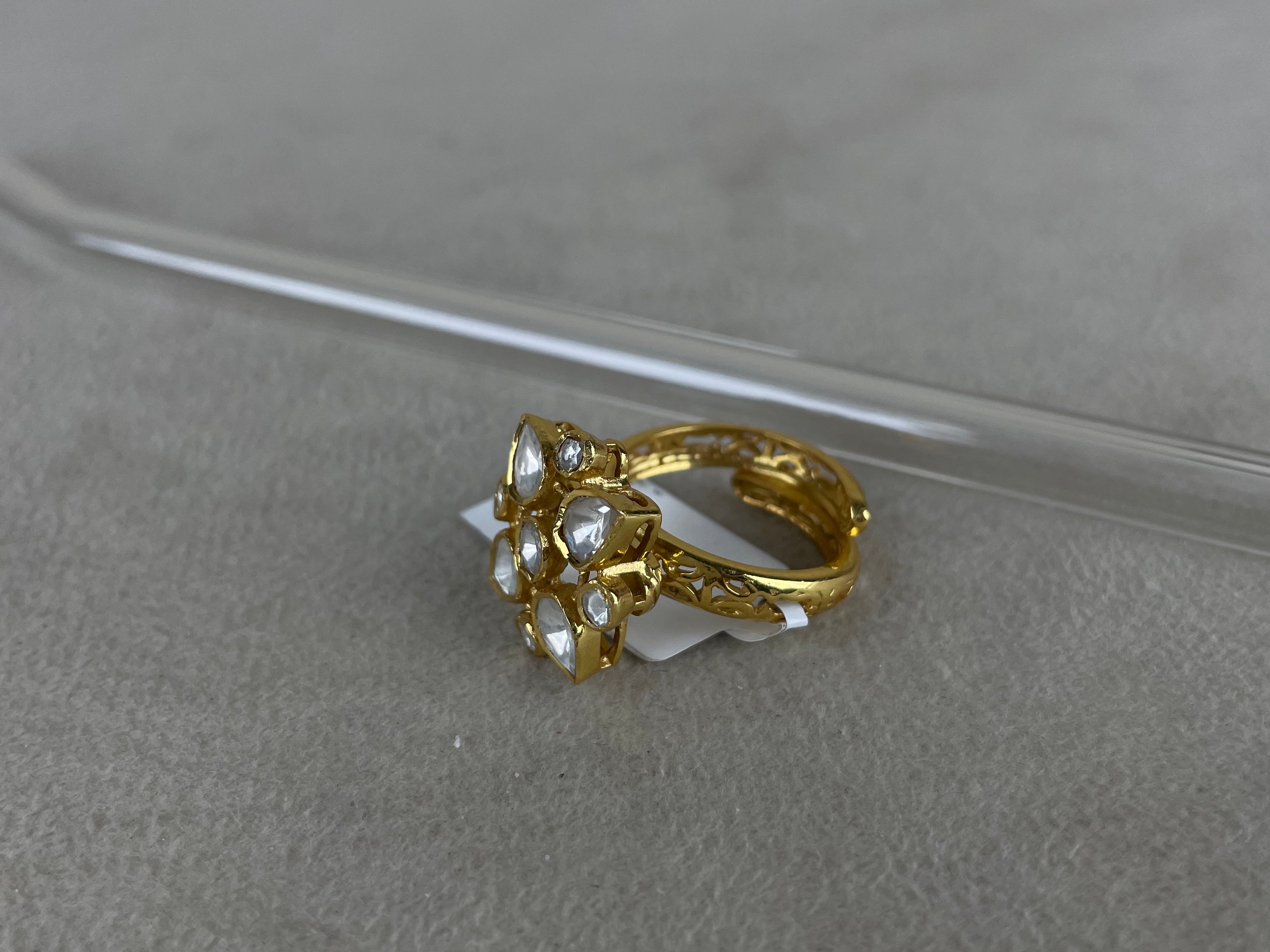 A gold-plated ring with a cluster of clear gemstones, resting on a textured gray surface with a clear glass rod in the background.