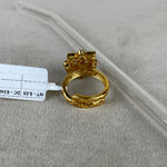 A gold-plated ring with a decorative top and textured band, shown on a neutral gray surface next to a clear plastic tube and a white tag.
