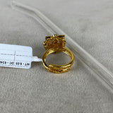 A gold-plated ring with a decorative top and textured band, shown on a neutral gray surface next to a clear plastic tube and a white tag.