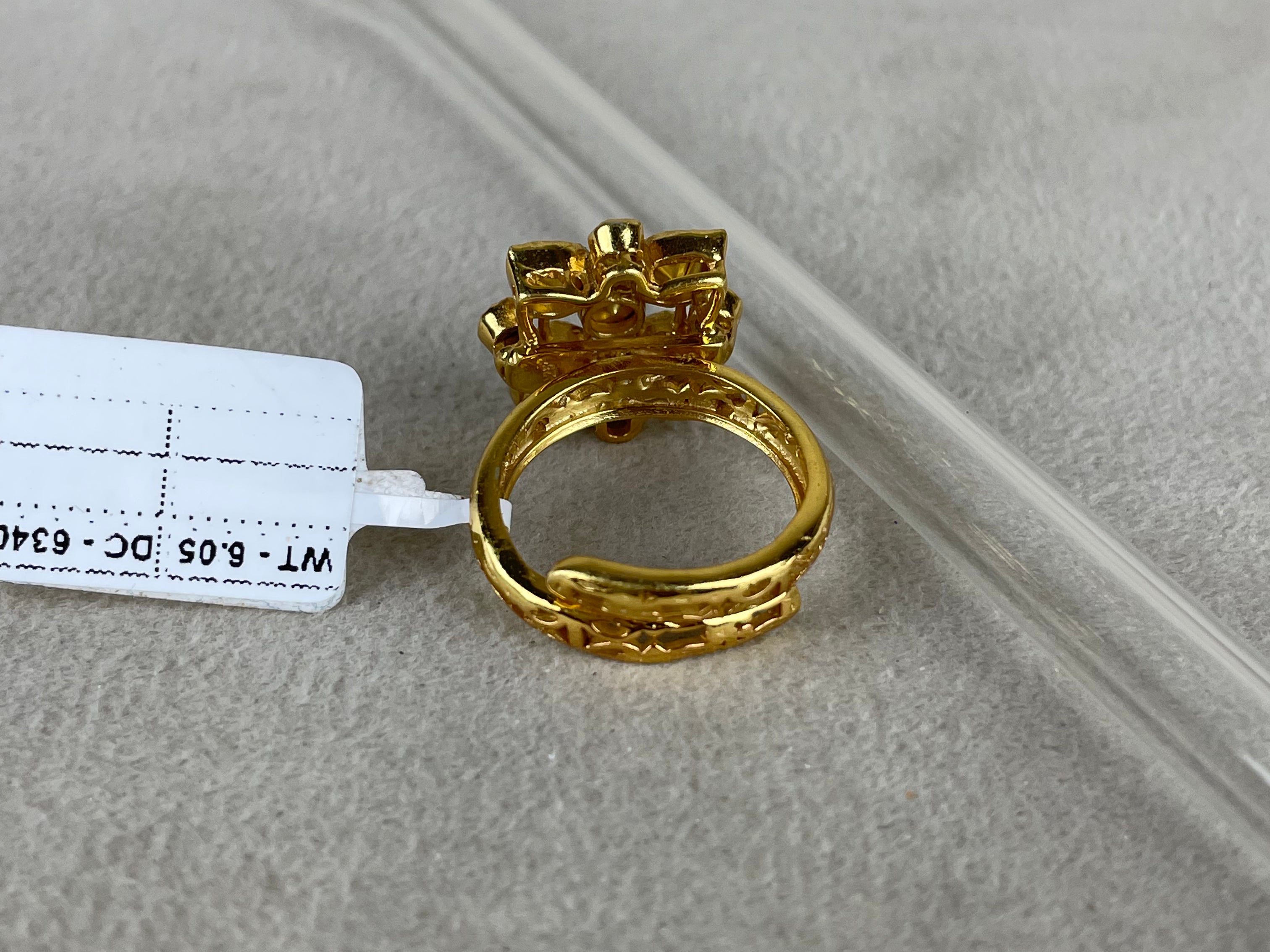 A gold-plated ring with a decorative top and textured band, shown on a neutral gray surface next to a clear plastic tube and a white tag.