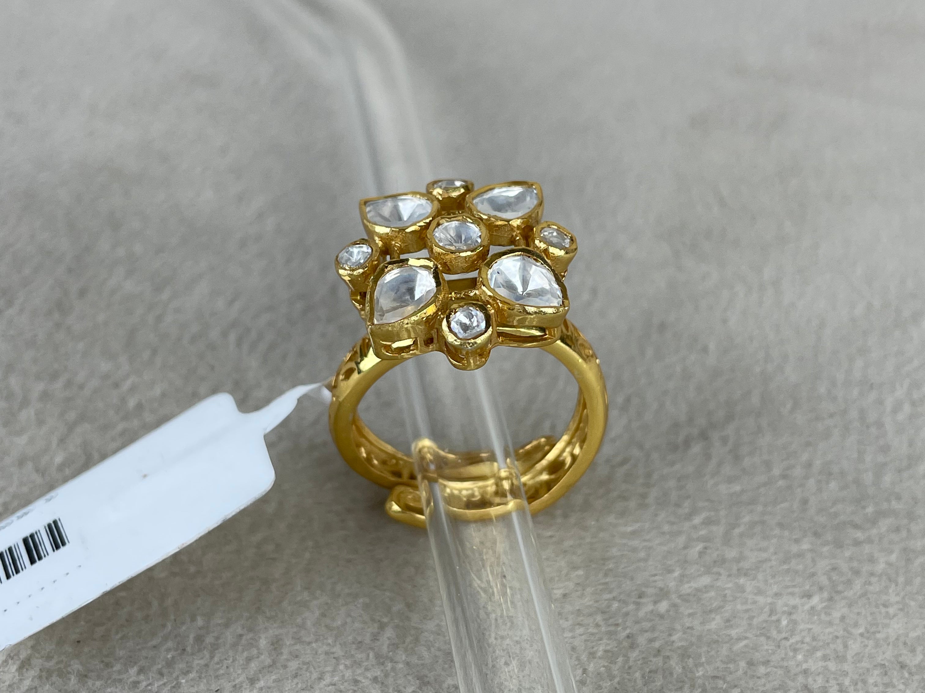 A gold ring with a floral cluster of clear stones, displayed on a clear plastic stand over a textured gray surface.