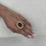 A gold-plated ring with a large green oval stone, a red rectangular stone, and white stones on a hand.