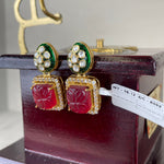 A pair of gold-plated Indian Kundan earrings with red carved stone panels, green enamel accents, and clear pavé rhinestones, displayed on a dark wooden box.