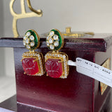 A pair of gold-plated Indian Kundan earrings with red carved stone panels, green enamel accents, and clear pavé rhinestones, displayed on a dark wooden box.