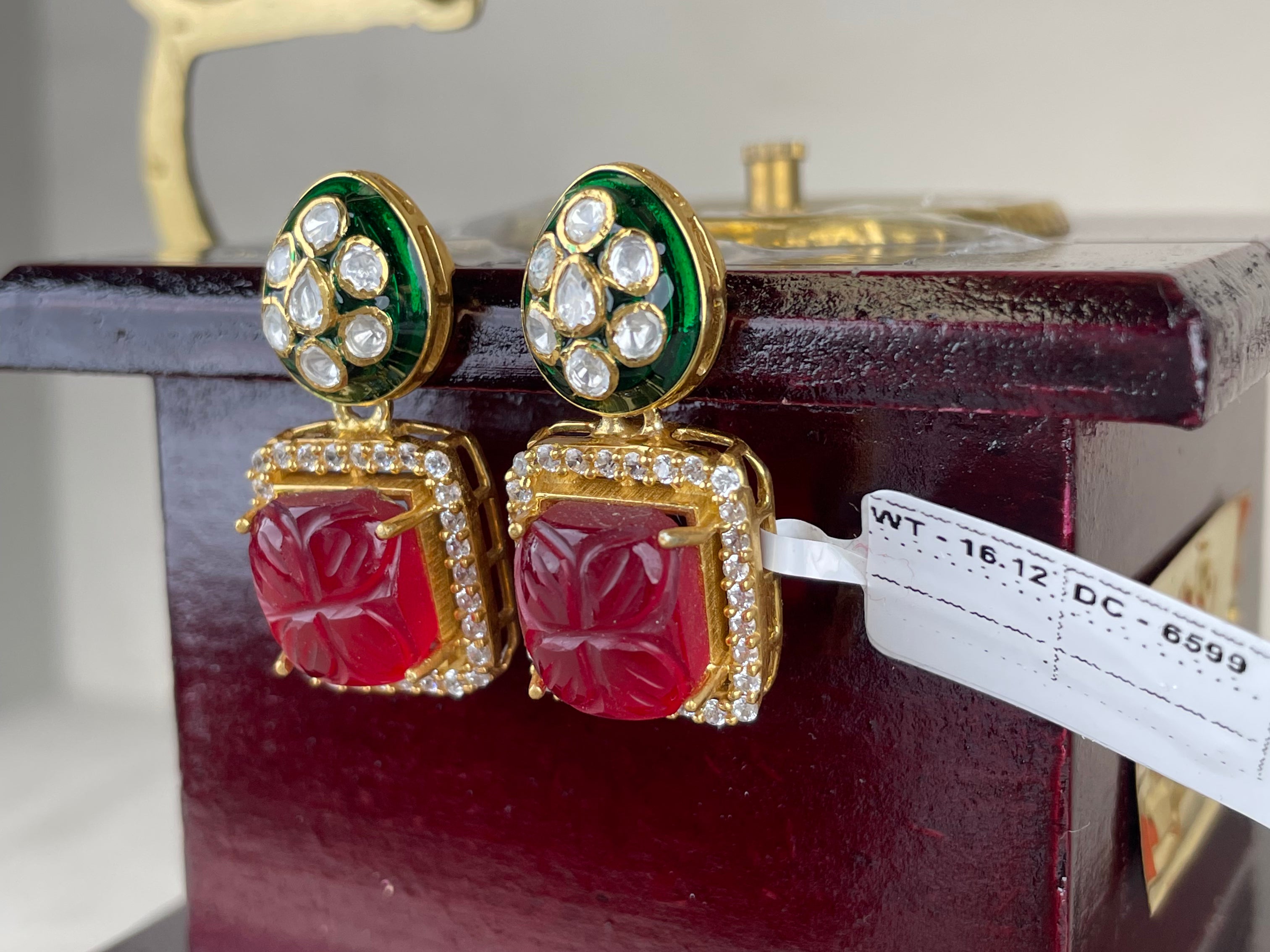 A pair of gold-plated Indian Kundan earrings with red carved stone panels, green enamel accents, and clear pavé rhinestones, displayed on a dark wooden box.