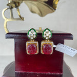 A pair of gold-tone statement earrings with green enamel, clear stones, and red carved floral drops, displayed on a dark red stand with a vintage gramophone prop in the background.