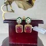 A pair of gold-tone statement earrings with green enamel, clear stones, and red carved floral drops, displayed on a dark red stand with a vintage gramophone prop in the background.