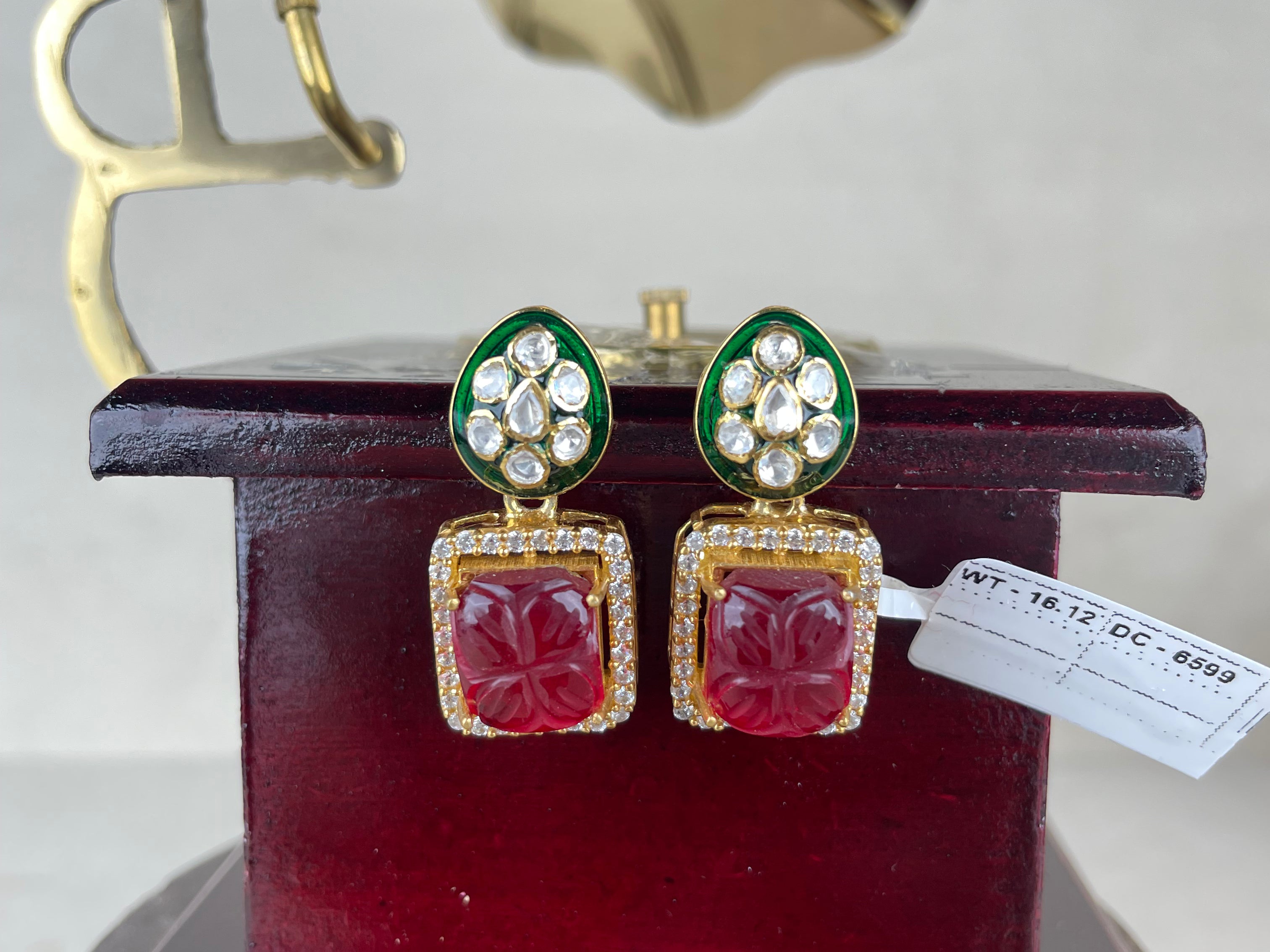 A pair of gold-tone statement earrings with green enamel, clear stones, and red carved floral drops, displayed on a dark red stand with a vintage gramophone prop in the background.
