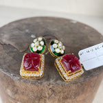 A pair of gold-plated Indian Kundan earrings featuring a large red stone in a square frame, bordered with small clear stones, and topped with green enamel and a cluster of clear stones, displayed on a rustic wooden block.