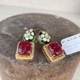 A pair of gold-plated Indian Kundan earrings featuring a large red stone in a square frame, bordered with small clear stones, and topped with green enamel and a cluster of clear stones, displayed on a rustic wooden block.