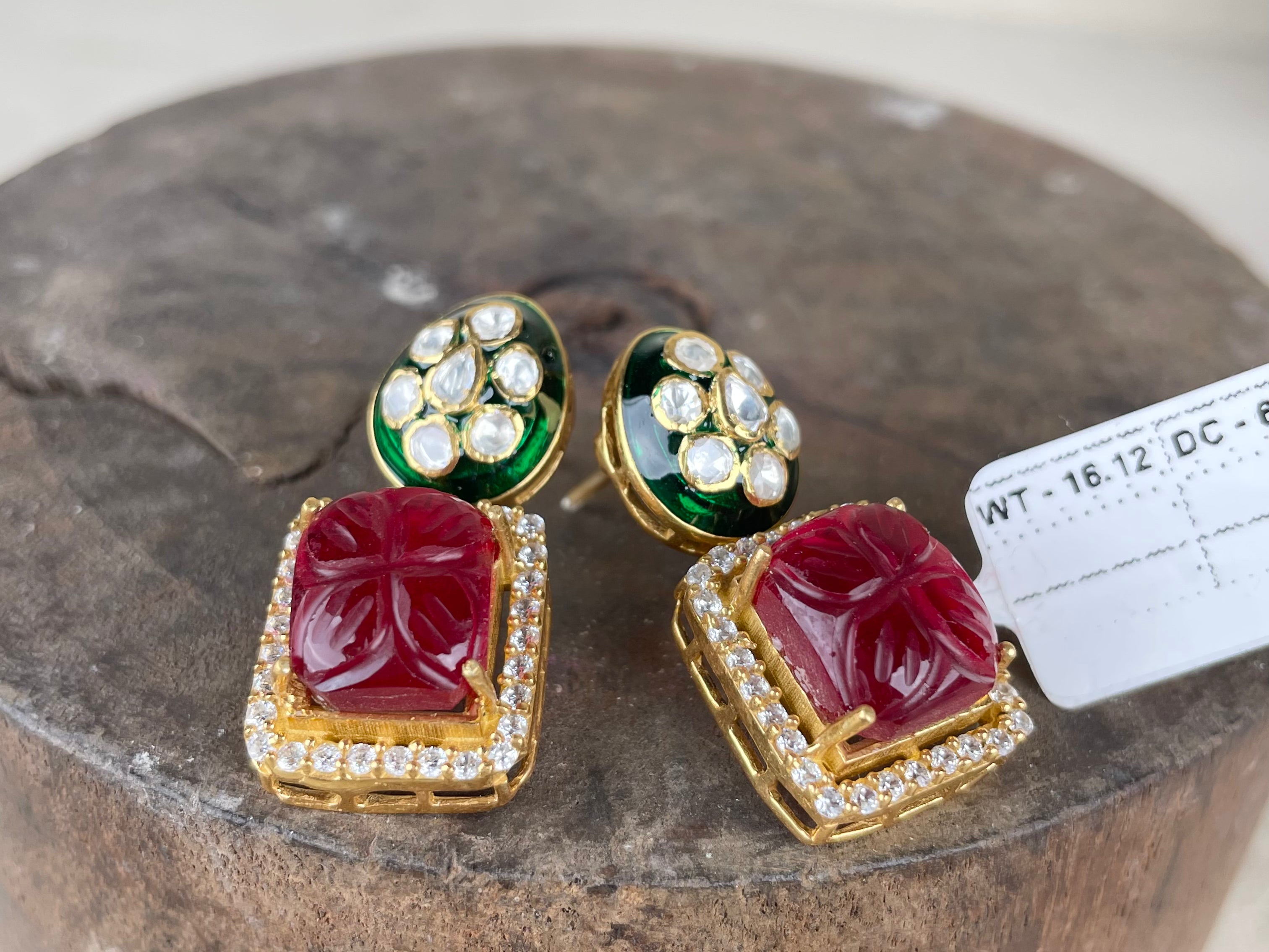 A pair of gold-plated Indian Kundan earrings featuring a large red stone in a square frame, bordered with small clear stones, and topped with green enamel and a cluster of clear stones, displayed on a rustic wooden block.