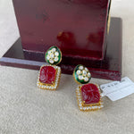 A pair of gold-plated Indian Kundan earrings with carved red stones and green enamel accents, displayed on a neutral surface in front of a dark wooden box.