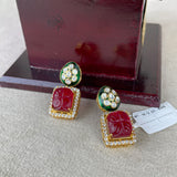 A pair of gold-plated Indian Kundan earrings with carved red stones and green enamel accents, displayed on a neutral surface in front of a dark wooden box.