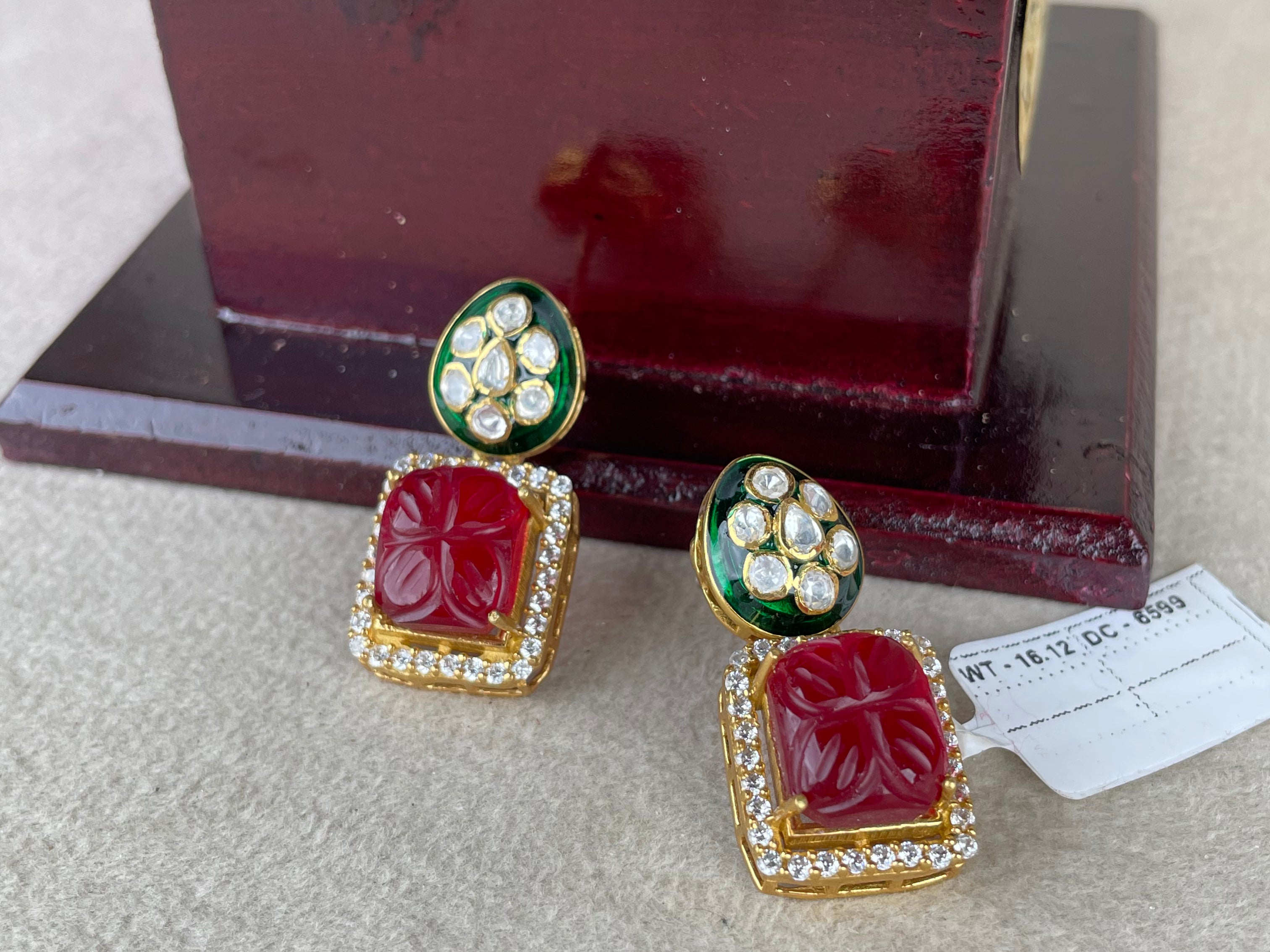 A pair of gold-plated Indian Kundan earrings with carved red stones and green enamel accents, displayed on a neutral surface in front of a dark wooden box.