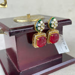 A pair of gold-plated drop earrings with red cabochon stones and green enamel accents, displayed on a dark wooden stand.