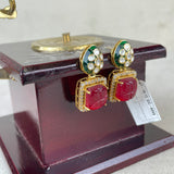 A pair of gold-plated drop earrings with red cabochon stones and green enamel accents, displayed on a dark wooden stand.