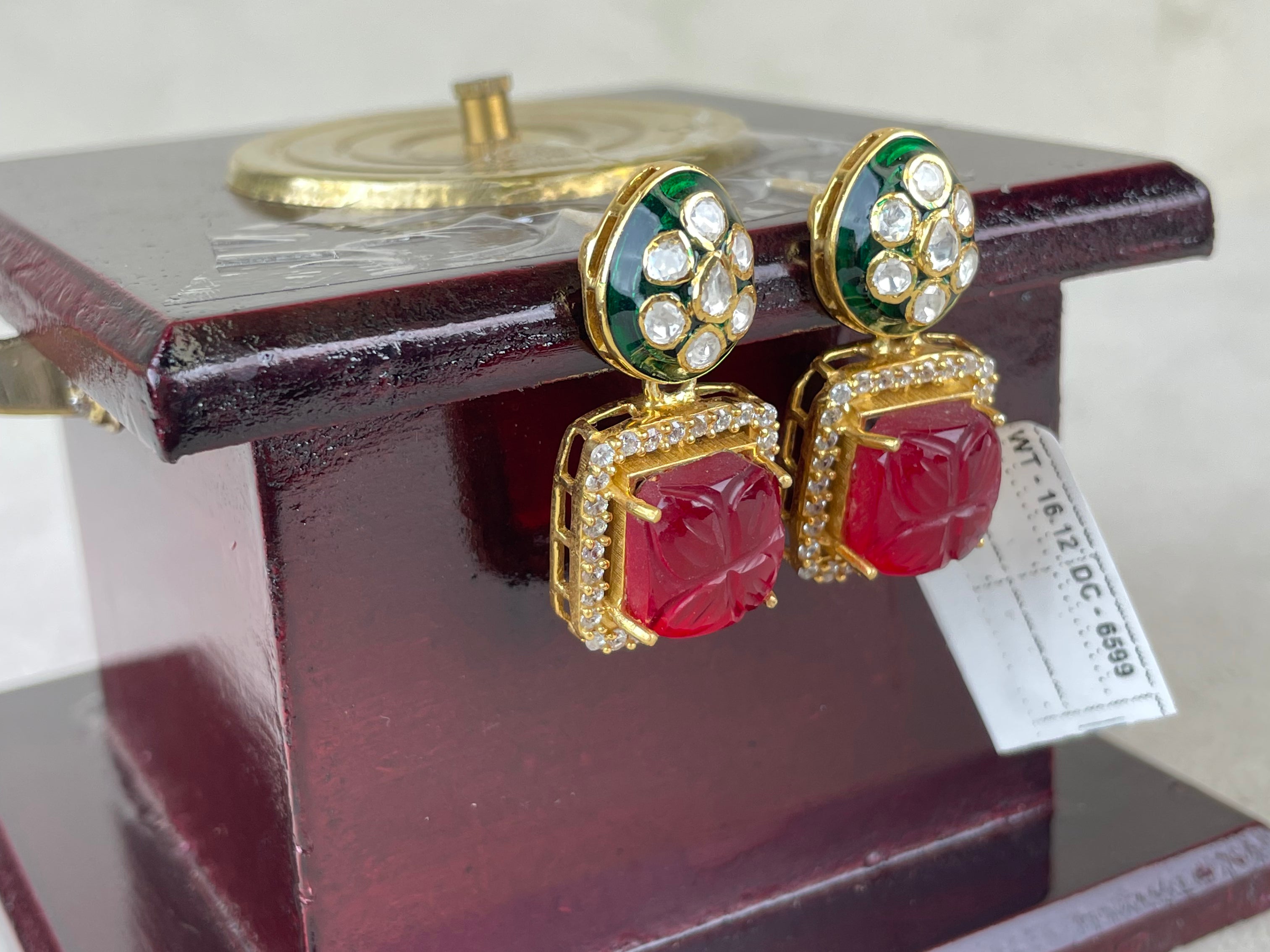 A pair of gold-plated drop earrings with red cabochon stones and green enamel accents, displayed on a dark wooden stand.