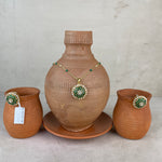 A terracotta clay set featuring a large decorative vase on a matching saucer, flanked by two smaller clay pots, with a gold-tone necklace and matching brooch displayed on the vase.