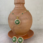 Gold and green jewelry set displayed on a terracotta vase, featuring a necklace with a round pendant and matching earrings with a white price tag attached.